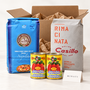 Ambient pizza-making gift set with one 1kg Dallagiovanna Tipo 00 flour bag, one 1kg Casillo semolina bag, two 400g Rega San Marzano tomato tins and a Minati card beside a plain postal carton. Illustrative arrangement.