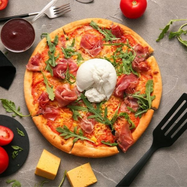 Pizza with prosciutto, arugula, and cheese on a gray surface with tomatoes and a spatula.