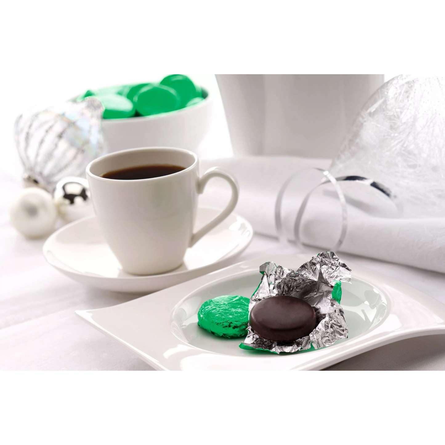 White cup of coffee with green and chocolate cookies on a white plate, with a white napkin and bowl in the background.